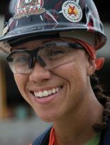 A young woman in construction helmet and goggles smiles at the camera